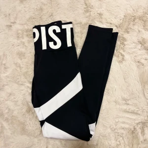 Svarta träningstights från Elly Pistol - Snygga svarta träningstights från Elly Pistol med vita diagonala ränder och textdetaljer i midjan. Perfekta för en sportig look eller träning. Tillverkade i ett stretchigt material för bästa komfort. Nya utan prislapp! 