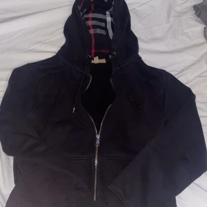 Svart hoodie från Burberry - Säljer en snygg svart hoodie från Burberry med dragkedja och broderad logga. Den har en klassisk rutig insida i huvan och dragsko. Perfekt för en stilren look. Inga defekter. Använd i typ 3 månader. Priset är inte hugget i stenen. INGA BYTEN