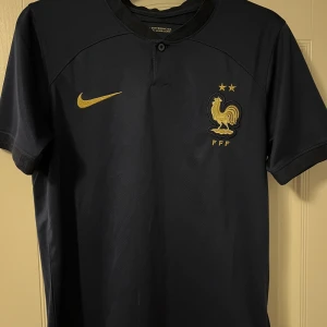 Frankrikes fotbollströja  - Frankrikes fotbolls tröja. Snygg mörkblå fotbollströja från Nike med korta ärmar. Tröjan har ett guldigt Nike-märke och en broderad tupp med två stjärnor på bröstet. Perfekt för fotbollsfans som vill visa sitt stöd med stil. 