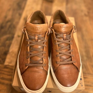 Bruna sneakers från Lloyd  - Snygga bruna sneakers i skinn från Lloyd med snörning och vit sula. Perfekta för en stilren look med en touch av elegans. Skorna har en diskret logga på sulan och en bekväm passform.