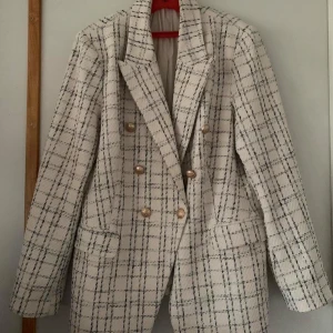 Vit rutig kavaj från Peppercorn - Snygg vit kavaj med svart rutmönster från Peppercorn. Kavajen har långa ärmar och guldknappar framtill samt på ärmsluten. Perfekt för en stilren look. Storlek L, skriv endast om du vill köpa.