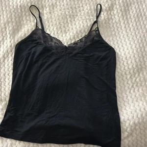 Svart linne med spetsdetaljer - Snyggt svart linne med tunna axelband och vacker spetsdetalj vid halsringningen. Perfekt för lager-på-lager eller som det är. Ett stilrent och mångsidigt plagg för garderoben.