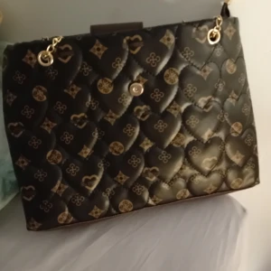 Svart quiltad handväska med hjärtmönster - Elegant svart handväska med quiltat hjärtmönster och guldfärgade detaljer. Väskan har en stilren design med en praktisk dragkedja och kedjehandtag. Perfekt för att ge en lyxig touch till din outfit.