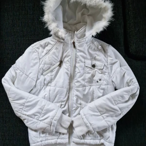 Vit dunjacka med fuskpäls från O&S - Säljer en snygg vit dunjacka från O&S med fuskpäls på huvan. Jackan stängs med dragkedja och har flera praktiska fickor med knappar. Även ribbade muddar och markerad midja. Finns en liten (obehandlad) fläck på baksidan av ena ärmen, se sista bilden. I övrigt är den i fint skick! 😊