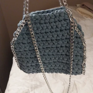 Blå virkad väska med kedja - Snygg blå virkad väska med silverfärgad kedja som axelrem. Perfekt för att ge en unik touch till din outfit. Väskan har en mjuk och mysig känsla tack vare det stickade materialet.