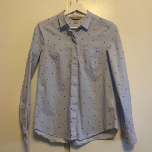 Blå randig skjorta från H&M - Säljer en stilren blå skjorta från H&M med smala ränder och små broderade detaljer. Skjortan har en klassisk krage och långa ärmar med knappar vid ärmsluten. Perfekt för en chic och avslappnad look.
