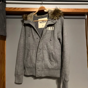 Fodrad jacka med päls - Säljer en grå jacka från Hollister med fuskpäls i luvan och broderad text '1922' på bröstet. Jackan har dragkedja och ribbade muddar. Perfekt för kyliga dagar.