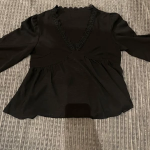Svart blus med spetsdetaljer - Elegant svart blus med v-ringning och spetsdetaljer längs halsringningen. Blusen har långa ärmar och en lätt utsvängd passform nedtill, vilket ger en fin siluett. Perfekt för en stilren look. Aldrig använd❤️