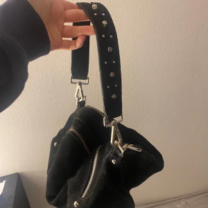 Svart axelväska med nitar - Snygg svart axelväska i mocka med silverfärgade dragkedjor och nitar på axelremmen. 