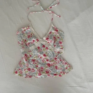 Blommig omlottopp med spetsdetaljer - Charmig blommig omlottopp med spetsdetaljer och knytband i nacken. Toppen har ett vackert mönster i rosa och grönt på en vit bakgrund. Perfekt för sommarens varma dagar.