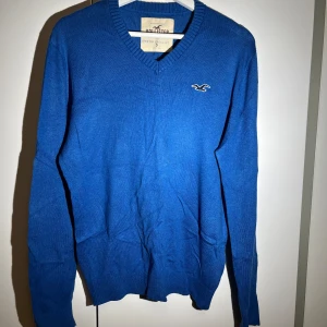 Blå tröja från Hollister - Säljer en snygg blå tröja från Hollister med v-ringning och ribbade kanter. Tröjan har en liten logga på bröstet och är perfekt för en stilren look. Passar bra till både jeans och chinos.