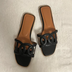 Svarta sandaler  - Stilrena svarta sandaler  med en elegant design. Sandalerna har en öppen tå och en dekorativ rem framtill. Perfekta för sommarens alla tillfällen.