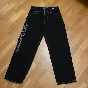 Jeans från Sweet SKTBS - Snygga svarta byxor från Sweet SKTBS med Street Skater broderad i vitt på benet. Byxorna har en rak och baggie passform. Köpta för 3 år sen oanvända, säljs eftersom det inte är min stil längre. Ny pris=900kr