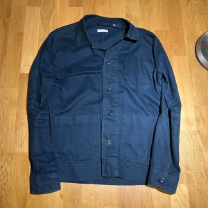 Mörkblå overshirt från Knowledge Cotton Apparel - Snygg mörkblå overshirt från Knowledge Cotton Apparel. Den har långa ärmar och knappar framtill. Perfekt för lager-på-lager-stil och passar bra till både vardag och mer avslappnade tillfällen. Storlek L passar M