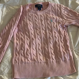 Rosa kabelstickad tröja från Ralph Lauren - Säljer en snygg rosa kabelstickad tröja från Ralph Lauren med det klassiska logotypbroderiet på bröstet. Tröjan har en rund halsringning och långa ärmar, perfekt för kyligare dagar. Passar både till jeans och kjol! Varan är i fint skick, barn strl 8-10 år 