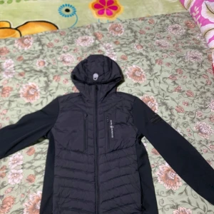 Svart jacka från Sail Racing - Snygg svart jacka från Sail Racing med luva och dragkedja. Jackan har en quiltad design och är perfekt för kyligare väder och är stilren.