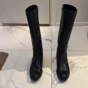 Svarta höga stövlar med dragkedja - Snygga svarta stövlar i skinn med dragkedja på sidan. Storlek 38. Perfekta för höst och vinter, dessa stövlar har en klassisk design med rund tå och låg klack. Äkta läder. Skorna är från ett italienskt märke.