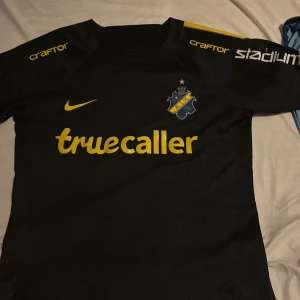 Svart AIK fotbollströja från Nike - Säljer en svart AIK fotbollströja från Nike med gula detaljer och Dri-FIT material. Tröjan har korta ärmar och sponsorer som Truecaller och SVEA tryckta i gult. Perfekt för AIK-fans som vill visa sitt stöd!