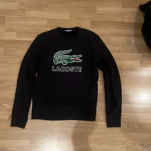 Svart sweatshirt från Lacoste - Säljer en svart sweatshirt från Lacoste med det ikoniska krokodilmärket på bröstet. Tröjan har långa ärmar och en klassisk passform. Perfekt för en stilren och avslappnad look.
