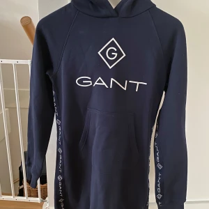Mörkblå hoodie från GANT - Säljer en snygg mörkblå hoodie från GANT med stor logga på bröstet och längs ärmarna. Tröjan har en praktisk magficka och en bekväm huva. Perfekt för en avslappnad stil. Man kan både använd den som hoodie och klänning beroende på❣️