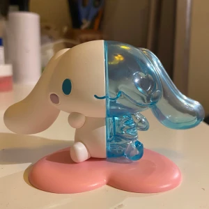 Cinnamoroll figur från Sanrio - Söt Cinnamoroll figur från Sanrio med en unik design där ena halvan är vit och den andra är genomskinligt blå. Figuren står på en rosa hjärtformad bas. Perfekt för samlare eller som dekoration!