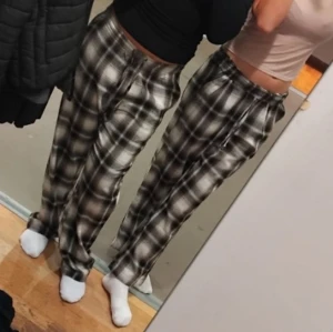Pyjamasbyxor i svart och grått - Säljer ett par bekväma och stiliga rutiga byxor i svart och grått. Byxorna har en loose passform och är perfekta för en avslappnad look. De har en elastisk midja med snörning för extra komfort.