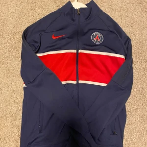 Blå träningsjacka från Nike - Säljer en blå träningsjacka från Nike med PSG-logga och rödvit rand över bröstet. Perfekt för sportiga aktiviteter. Dri-FIT-material för optimal komfort. Storlek L i barnstorlek.