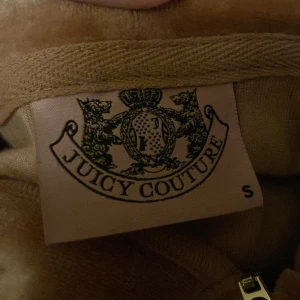 Brun velourjacka från Juicy Couture - Säljer en brun velourjacka från Juicy Couture med glittrande detaljer och dragkedja. Jackan har en huva och är dekorerad med strass på ryggen och huvan. Perfekt för en glamorös look!