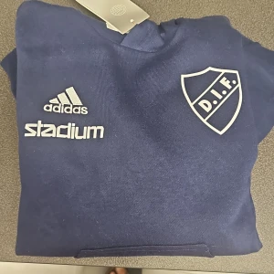 Blå hoodie från Adidas med DIF-logga - Säljer en blå hoodie från Adidas med DIF-logga och Stadium-märke. Tröjan har en klassisk känguruficka och är perfekt för sportiga tillfällen. Den är i storlek 140 och har en bekväm passform med långa ärmar.