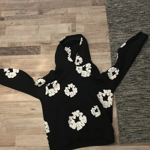Svart hoodie med blommönster från Denim Tears - Säljer en cool svart hoodie från Denim Tears med ett unikt vitt blommönster. Perfekt för att sticka ut i mängden. Hoodien har en klassisk passform och är tillverkad i mjukt material för extra komfort.