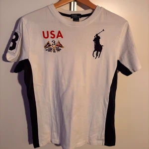 Vit t-shirt med USA-tryck från Polo Ralph Lauren - Snygg vit t-shirt från Polo Ralph Lauren med rött USA-tryck och den klassiska loggan framtill. T-shirten har korta ärmar och svarta sidopaneler. Perfekt plagg till vardags eller finare tillställningar. Använd ett fåtal gånger. Storlek L i barn (14-16) men passar perfekt för S i vuxen storlek!