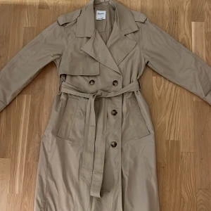 Beige trenchcoat från Bershka - Snygg beige trenchcoat från Bershka med klassisk design. Jackan har knappar och ett bälte i midjan för en stilren look. Perfekt för vår och höst.