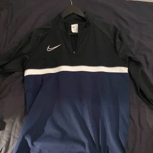 Svart och blå långärmad tröja från Nike - Säljer en svart och blå långärmad tröja från Nike med dragkedja och vit rand över bröstet. Perfekt för träning med Dri-FIT material som håller dig torr och bekväm. Nike-logga på bröstet ger en sportig look. Kom dm för fler frågor!