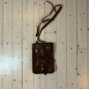 Brun axelväska i skinn för kartor eller iPads :) - Snygg brun axelväska i skinn med justerbar axelrem och spänndetaljer. Det är ursprungligen en militär kartväska för bära kartor. Perfekt för att bära din iPad i med både regnskydd å mjuk god vaddering med stil. Väskan har en praktisk design med flera fack och stängs med spänne i två riktnkngar. Perfekt när man ska ta en tur runt stan på en vespa eller liknande retro utflykt!