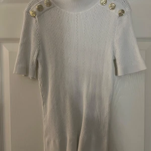 Vit tröja från Amisu - Säljer en stilren vit ribbad tröja från Amisu med korta ärmar och  guldknappar på axlarna. Perfekt för en elegant look. Tröjan är i storlek M.