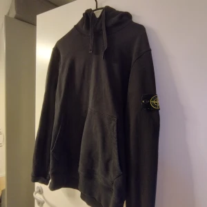 Svart hoodie från Stone Island - Säljer en svart hoodie från Stone Island med klassisk logga på ärmen. Tröjan har en bekväm passform med långa ärmar och en praktisk känguruficka framtill. Perfekt för en avslappnad stil.