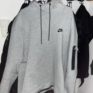 Grå nike tech hoodie - Säljer en grå hoodie från Nike med svart logga på bröstet. Den har en praktisk ficka med dragkedja på ärmen och justerbar huva med snören. Perfekt för en avslappnad stil. Storlek xl