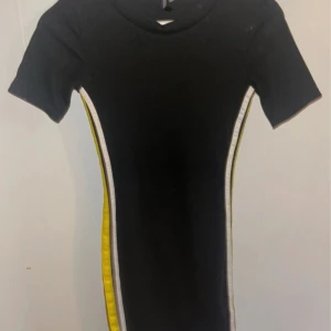 Svart klänning med gula ränder - Snygg svart t-shirt klänning med korta ärmar och gula ränder längs sidorna. Perfekt för en casual look med en sportig touch. Klänningen är figurnära och kort i modellen.