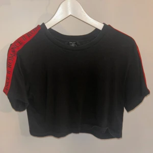 Svart croppad t-shirt med röd textdetalj - Snygg svart croppad t-shirt från Forever 21 med röd textdetalj längs ärmen. Perfekt för en avslappnad stil med en modern touch.