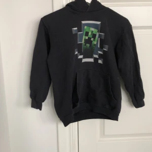 Svart hoodie med Minecraft-motiv - Cool svart hoodie med ett stort Minecraft Creeper-tryck på framsidan. Perfekt för gaming-fans! Tröjan har en klassisk passform med långa ärmar och en praktisk ficka framtill.