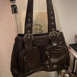 Brun axelväska från Franco Rino - Snygg brun axelväska i skinn från Fanco Rino med praktiska fickor framtill. Väskan har justerbara axelremmar med metallringar och flera nitar för en cool look. Perfekt för att bära med sig det viktigaste med stil.