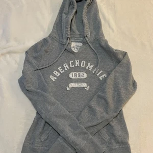 Grå hoodie från Abercrombie & Fitch - Säljer en stilren grå hoodie från Abercrombie & Fitch med tryck framtill. Tröjan har en klassisk design med huva och långa ärmar, perfekt för en avslappnad look. Passar utmärkt till vardags.