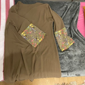 Brun abaya  med blommiga ärmslut - Snygg brun abaya med långa ärmar och färgglada blommiga ärmslut. Perfekt för en bohemisk look med en touch av färg. Klänningen har en avslappnad passform och är tillverkad i ett mjukt material.