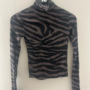 Zebramönstrad polotröja - Snygg zebramönstrad polotröja i svart och grått. Tröjan har en tight passform och långa ärmar, perfekt för en trendig look. Den höga kragen ger en extra stilpoäng.