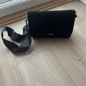 Svart axelväska från NA-KD - Snygg svart axelväska från NA-KD med justerbar axelrem och stilren design. Väskan har en praktisk dragkedja på toppen för säker förvaring. Skriv gärna vid frågor, pris kan diskuteras.