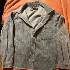 Grå manchesterjacka från Zara - Säljer en stilren grå manchesterjacka från Zara. Jackan har en klassisk krage, knappar framtill och en bröstficka. Perfekt för en avslappnad look.