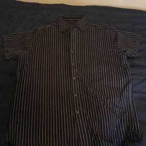 Svart och vit randig skjorta från Atlant - Snygg svart skjorta med vita ränder från Atlant. Skjortan är kortärmad och har knappar framtill. Perfekt för en stilren look. Storlek M, material 100% bomull. 