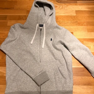 Grå hoodie från Polo Ralph Lauren - Säljer en stilren grå hoodie från Polo Ralph Lauren med dragkedja och justerbar huva. Den har en broderad logotyp på bröstet och är tillverkad i mjukt bomullsmaterial. Perfekt för en avslappnad look.
