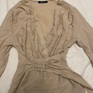 Beige omlottopp med bälte från Gina Tricot - Säljer en snygg beige omlottopp från Gina Tricot. Toppen har långa ärmar och ett matchande bälte i midjan som ger en fin siluett. Perfekt för både vardag och fest!