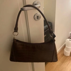 Brun axelväska från Bik Bok - Säljer en snygg brun axelväska från Bik Bok i mycket bra skick. Väskan har ett croco-liknande mönster och en praktisk dragkedja upptill. Perfekt för både vardag och fest! 👜